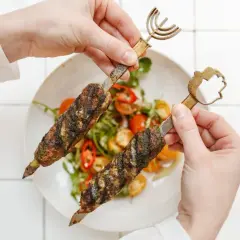 Dwie ręce trzymają szaszłyki z mięsem mielonym i falafelami na talerzu z sałatką.