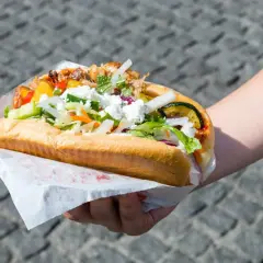 Ręka trzymająca dużą bułkę z mięsem, warzywami i serem feta.