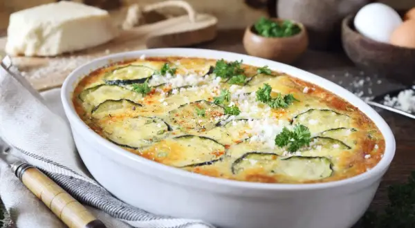Zapiekanka z cukinii w białym naczyniu, ozdobiona natką pietruszki i parmezanem.