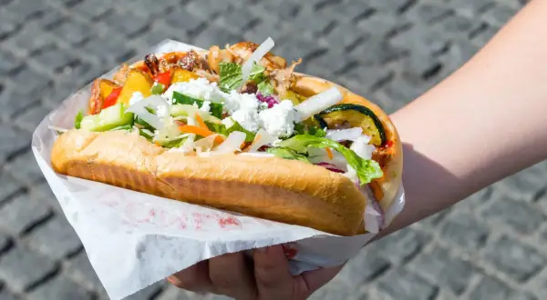 Ręka trzymająca dużą bułkę z mięsem, warzywami i serem feta.