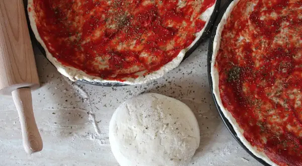 Dwa surowe ciasta na pizzę z sosem pomidorowym i ziołami, wałek i kulka ciasta.