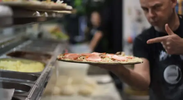 Mężczyzna w fartuchu pokazuje surową pizzę z szynką i pieczarkami.