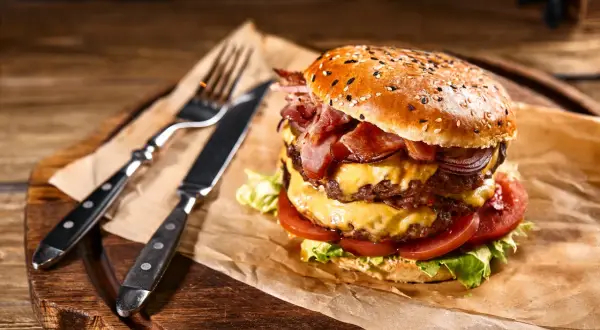 Soczysty burger z podwójną wołowiną, serem, bekonem i warzywami na desce z widelcem i nożem.